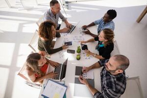 Sechs Erwachsene, die gemeinsam mit ihren Laptops, Tablets und Unterlagen an einem Tisch sitzen und sich angeregt unterhalten.