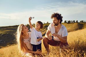 Dreiköpfige Familie bestehend aus Mutter, Vater und Sohn auf einem Feld. Der Sohn bläst Seifenblasen, während Vater und Mutter ihn anschauen.