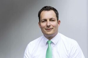 Mann mit kurzen braunen Haaren, lächelnd. Trägt ein weißes Hemd und eine hellgrüne Krawatte. Der Hintergrund ist weiß.