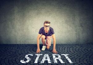 Junger Mann in Alltagskleidung der sich in der klassischen Startposition für ein Wettrennen befindet. Er kniet auf Kopfsteinpflaster, vor ihm auf dem Boden der Text "START".